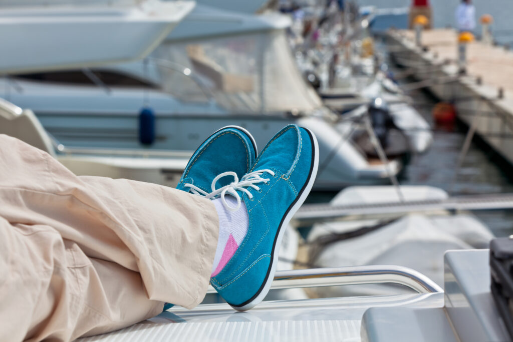 Someone's wearing beige pants, white socks with pink detail, and blue shoes, relaxing with their feet up on a docked boat. Other boats and a marina are visible in the background.