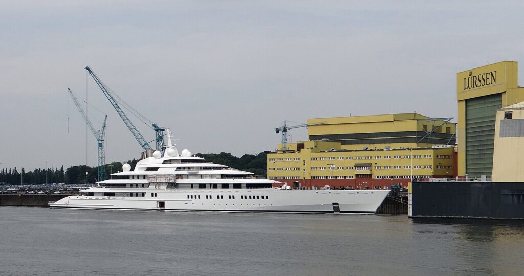 A large white luxury yacht, the Azzam, is docked beside the Lürssen shipyard, with cranes and industrial buildings in the background.