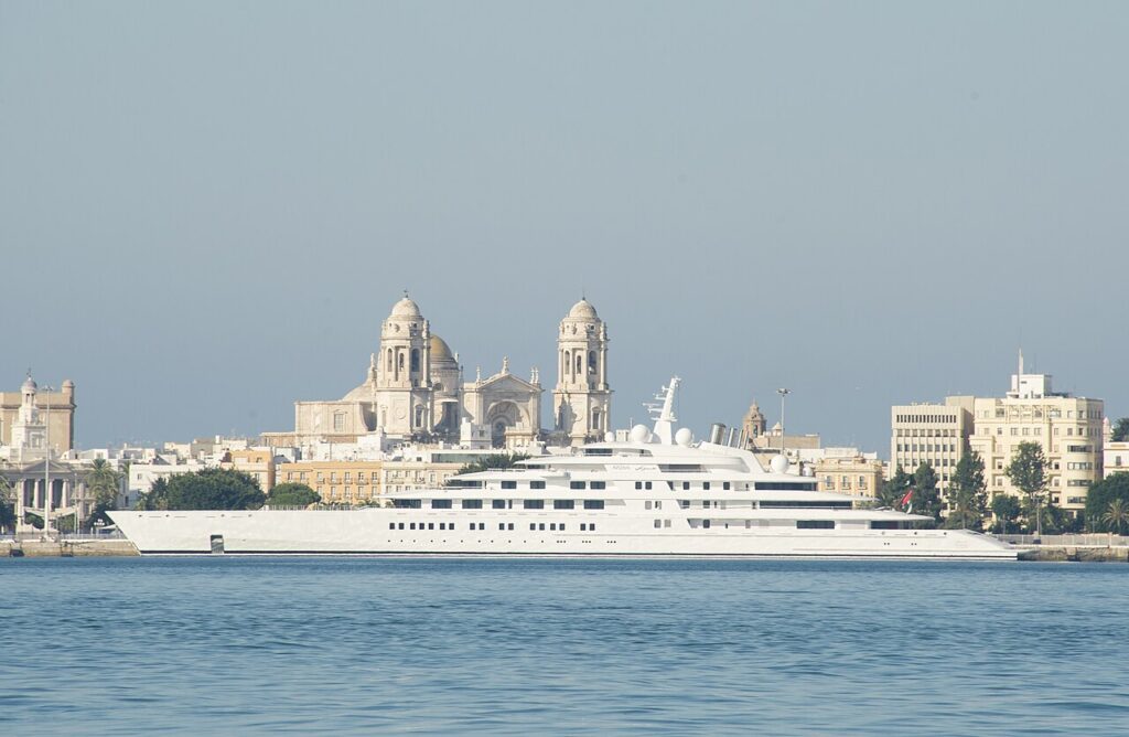 A large white yacht's anchored in a harbor, with historic buildings and a Cathedral visible in the background under a clear sky.