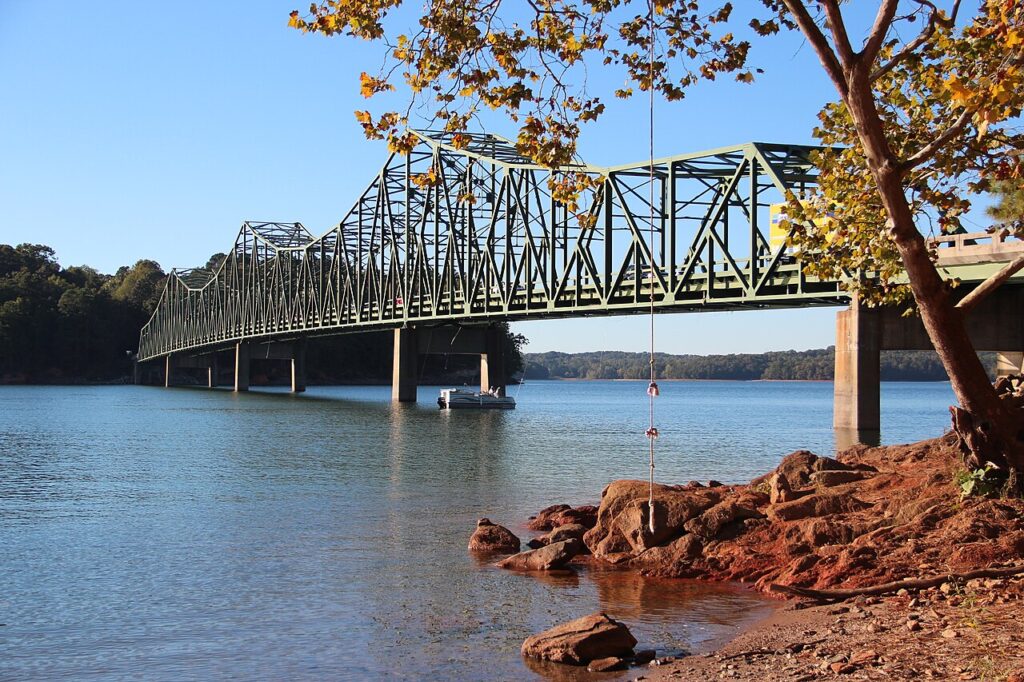 A green steel truss bridge spans a wide river—one of the best boating lakes—with a boat below it. Tree branches and a rocky, red shoreline fill the foreground.