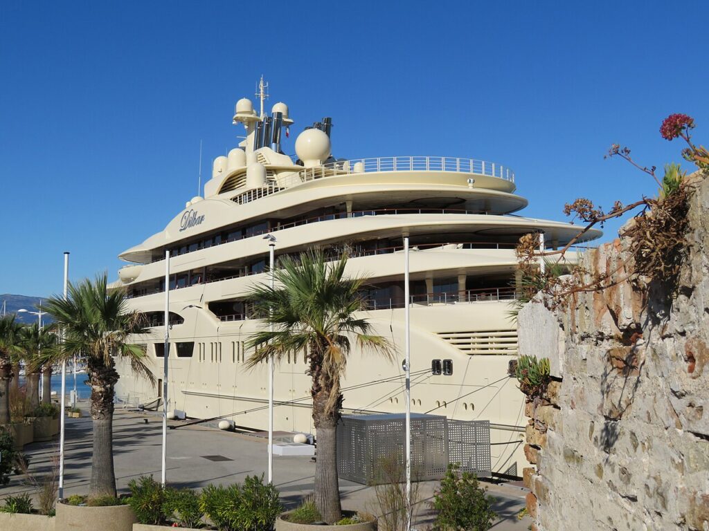 There's a large beige luxury yacht, the Dilbar, docked at a marina lined with Palm Trees, and there's a clear blue sky overhead.