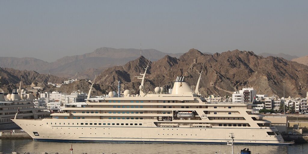 A large, white luxury yacht, the Fulk Al Salamah, is docked in a harbor with rocky mountains and city buildings in the background under a clear sky.