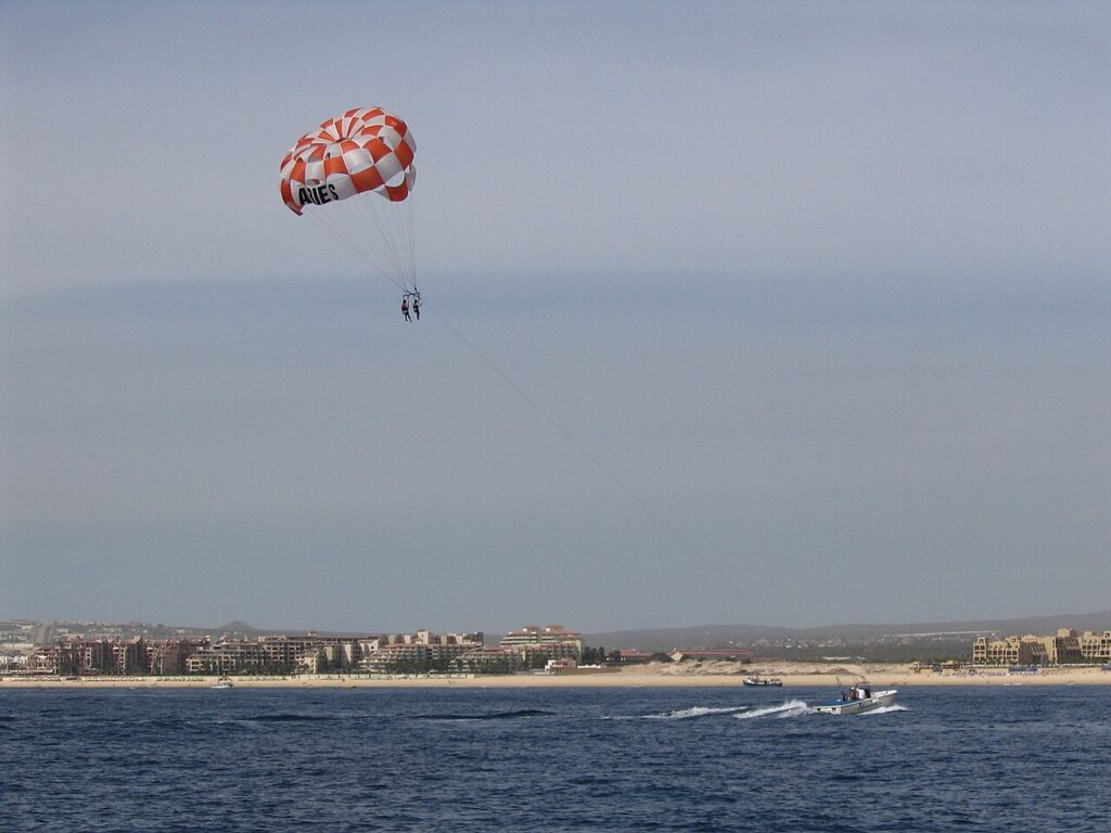 Two people are parasailing over the ocean, attached to a boat by a long rope. The city shoreline and buildings are visible in the background.