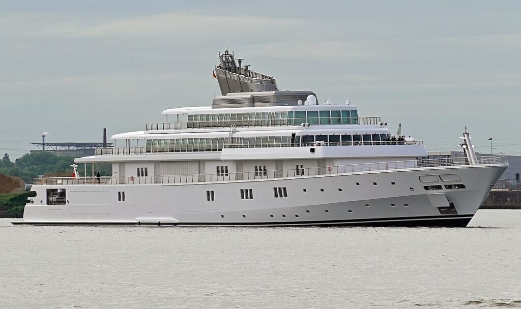 A large, modern white motor yacht with multiple decks is cruising on a body of water near an industrial shoreline.
